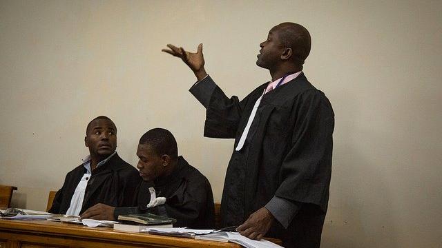 A lawyer wearing a black robe and a pink business shirt standing up in court addressing the room.