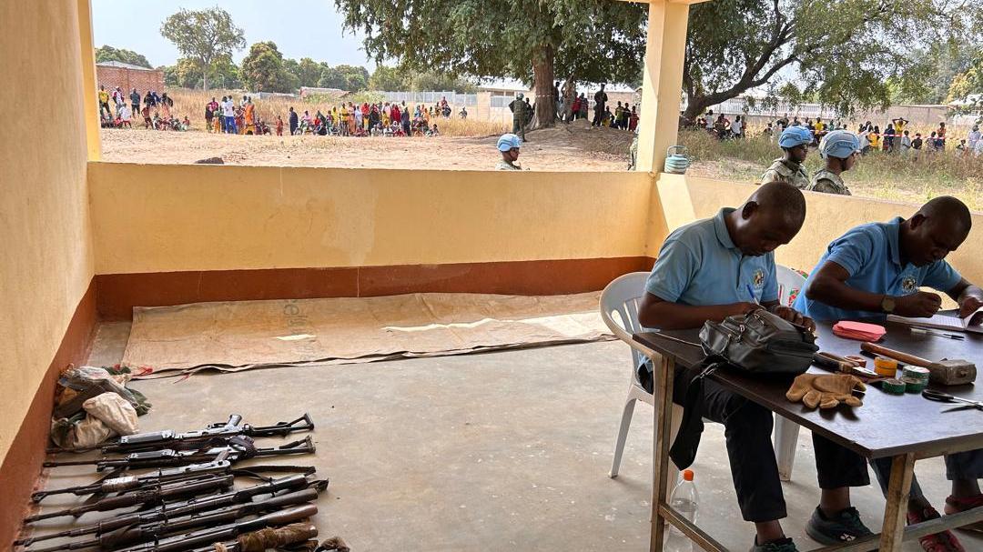 Two individuals seated at a table inside a covered area, working with documents and items. Several rifles are laid out on the floor nearby. Outside, a group of people and uniformed personnel can be seen in an open area with trees.