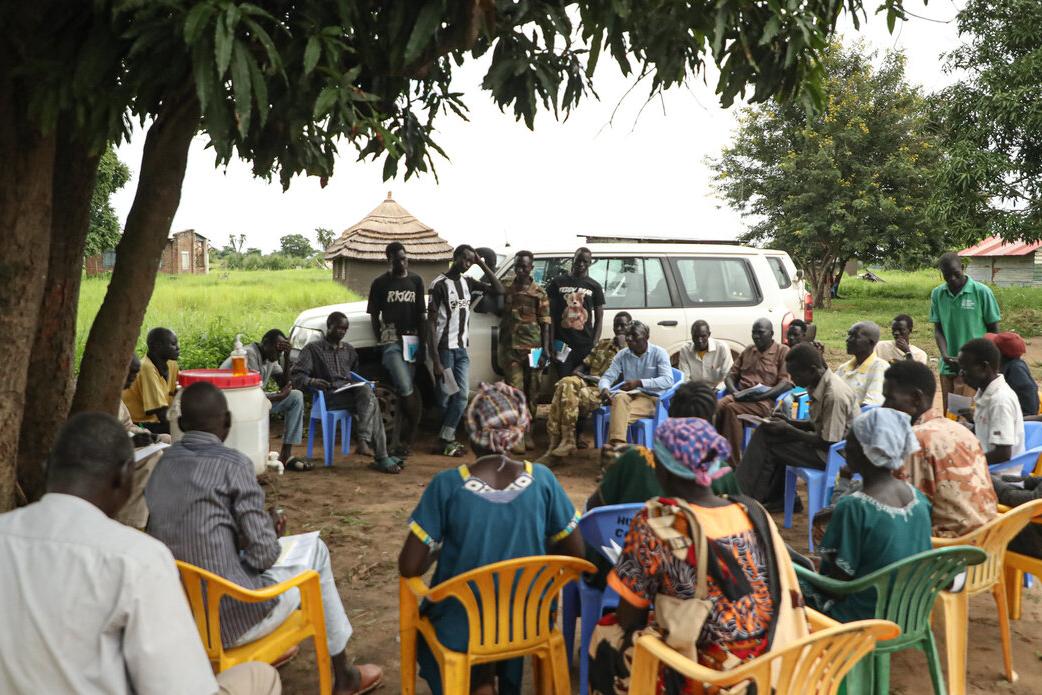 A visiting UNMISS team engaged with local communities in Gondokoro, where land conflicts between farmers and cattle herders from elsewhere have led to tensions and insecurity. Community meeting held outdoors under a tree, with people seated in a circle on plastic chairs and several individuals standing near a white vehicle in the background.