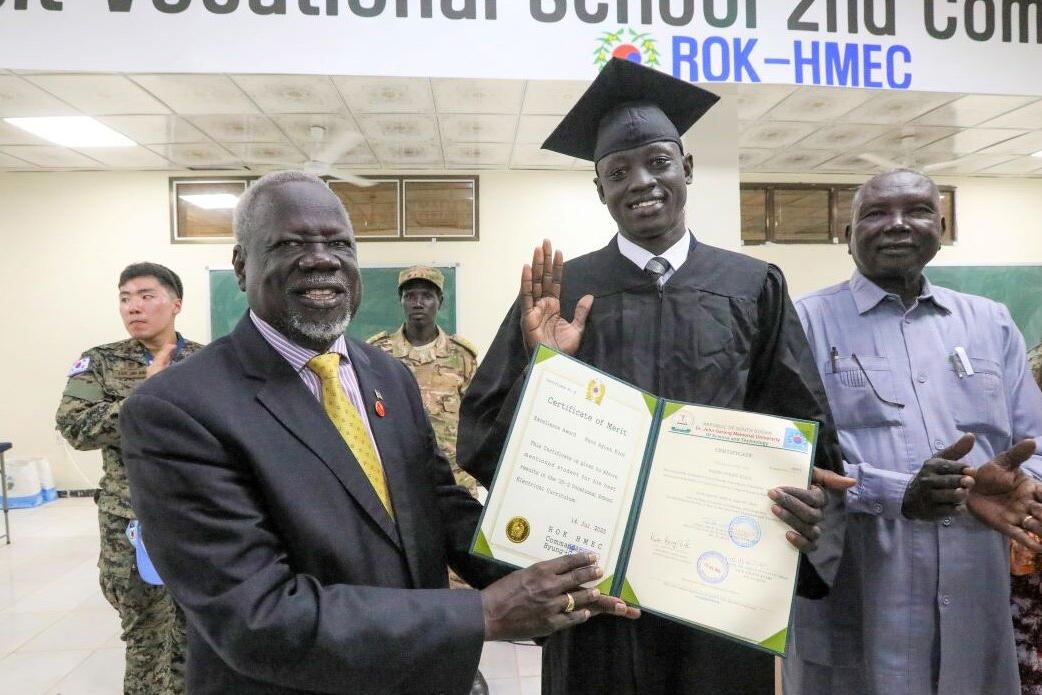 Youth in South Sudan often have difficulties finding learning opportunities and jobs. In Bor, peacekeepers from South Korea have conducted (Bor) 3-month-long vocational trainings for young, and now optimistic, women and men. In Akobo, UNMISS has funded a similar initiative. Graduation ceremony indoors with a person in a cap and gown holding a certificate, surrounded by others, with a banner for a vocational school visible above.