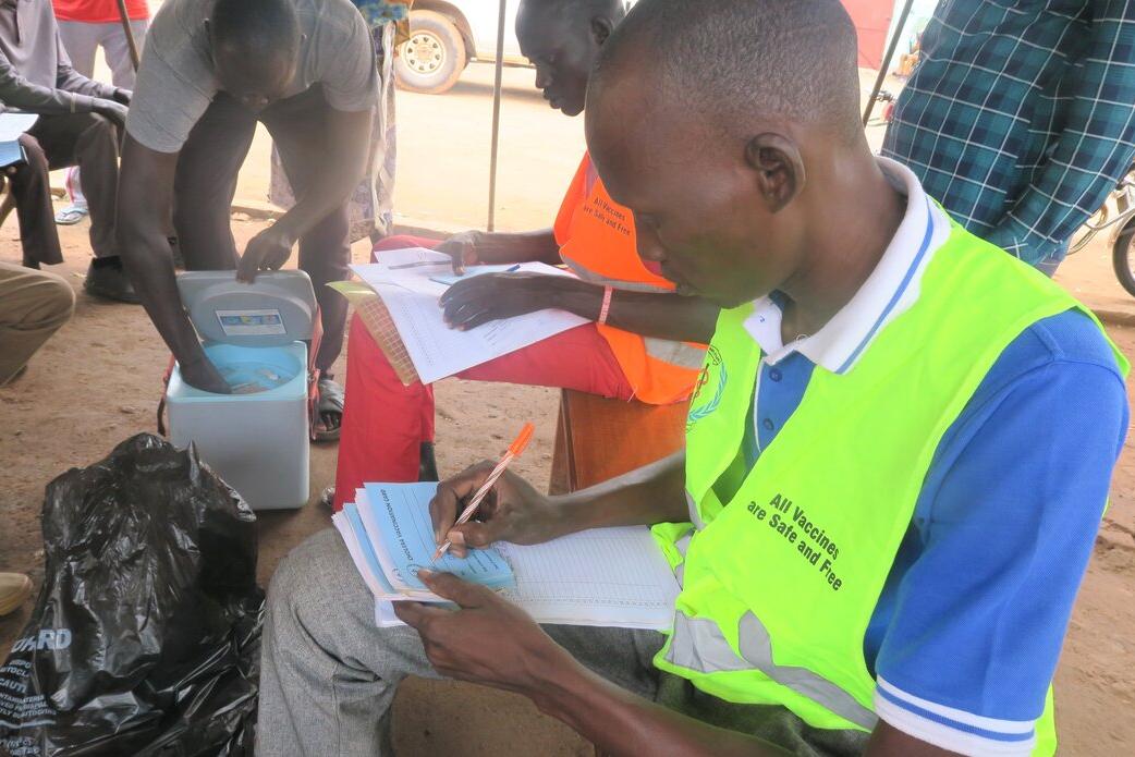 With a capacity of 150 inmates, Aweil Central Prison in Northern Bahr El Ghazal currently houses over 700 prisoners in crowded, unhygienic conditions. With cholera running rampant in the region, UNMISS advocated with local authorities and the World Health Organization to vaccinate all prisoners and their guards. Health worker wearing a bright vest writes in a register during a vaccination campaign, with a cooler box and other people seated nearby.