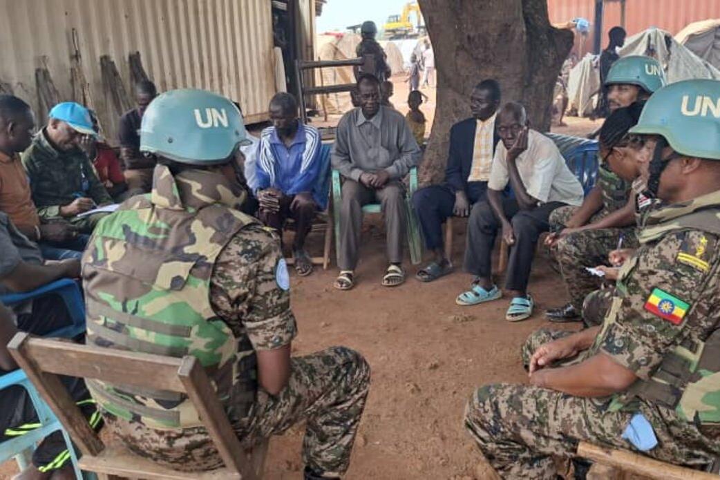 UN peacekeepers in camouflage uniforms and blue helmets sit in a circle outdoors, engaging in discussion with local community members near tents and a corrugated structure