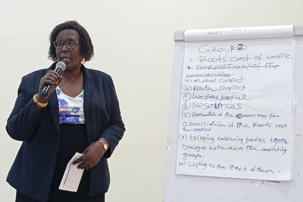 Person holding a microphone and standing beside a flip chart with handwritten notes on conflict causes and committee formation.